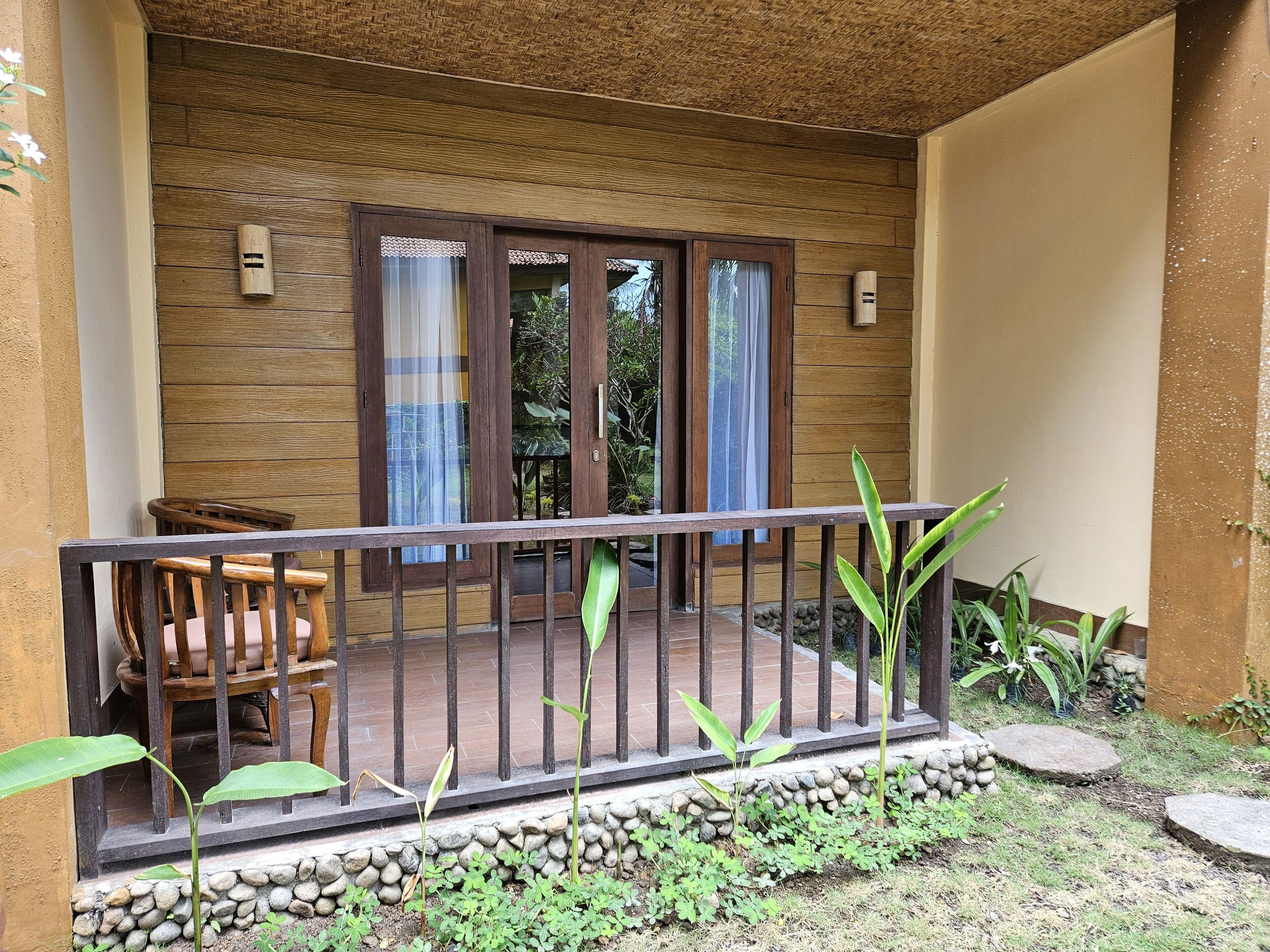 A wooden balcony with a railing, a chair, and a small garden area with plants and a stone wall.