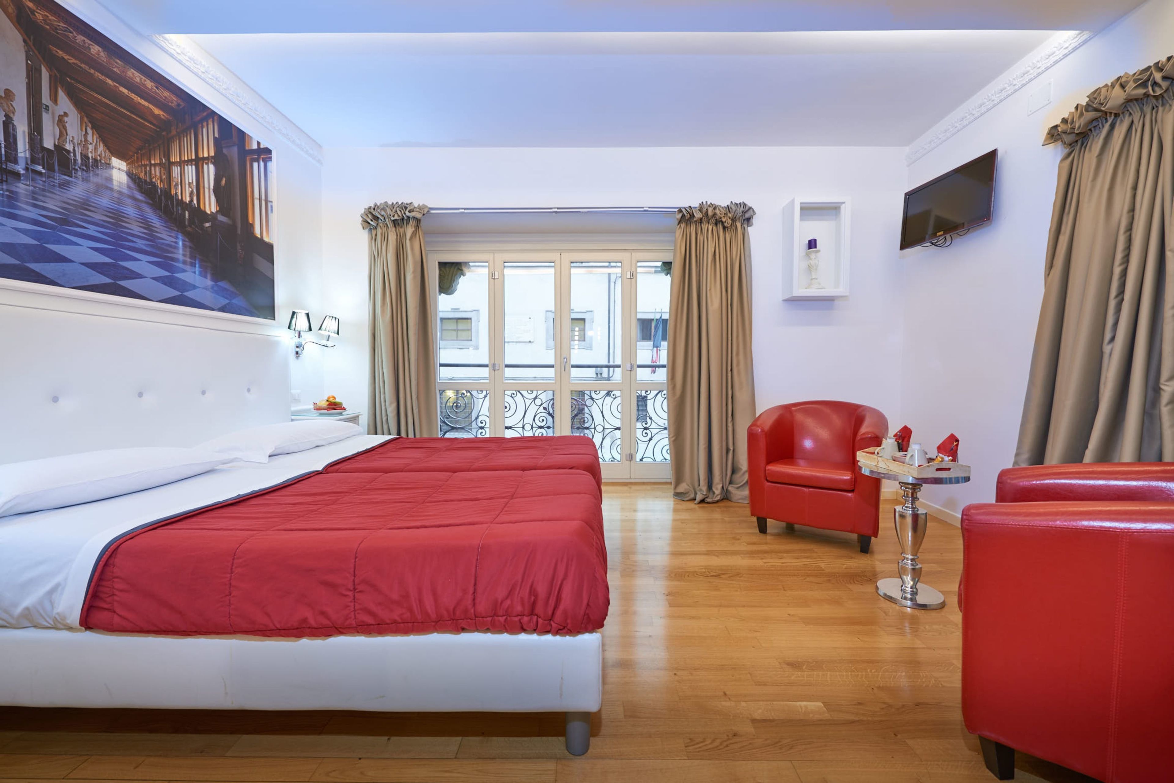 Habitación de hotel con una cama grande, un sillón rojo, una mesita y vistas a la ciudad a través de un ventanal amplio.