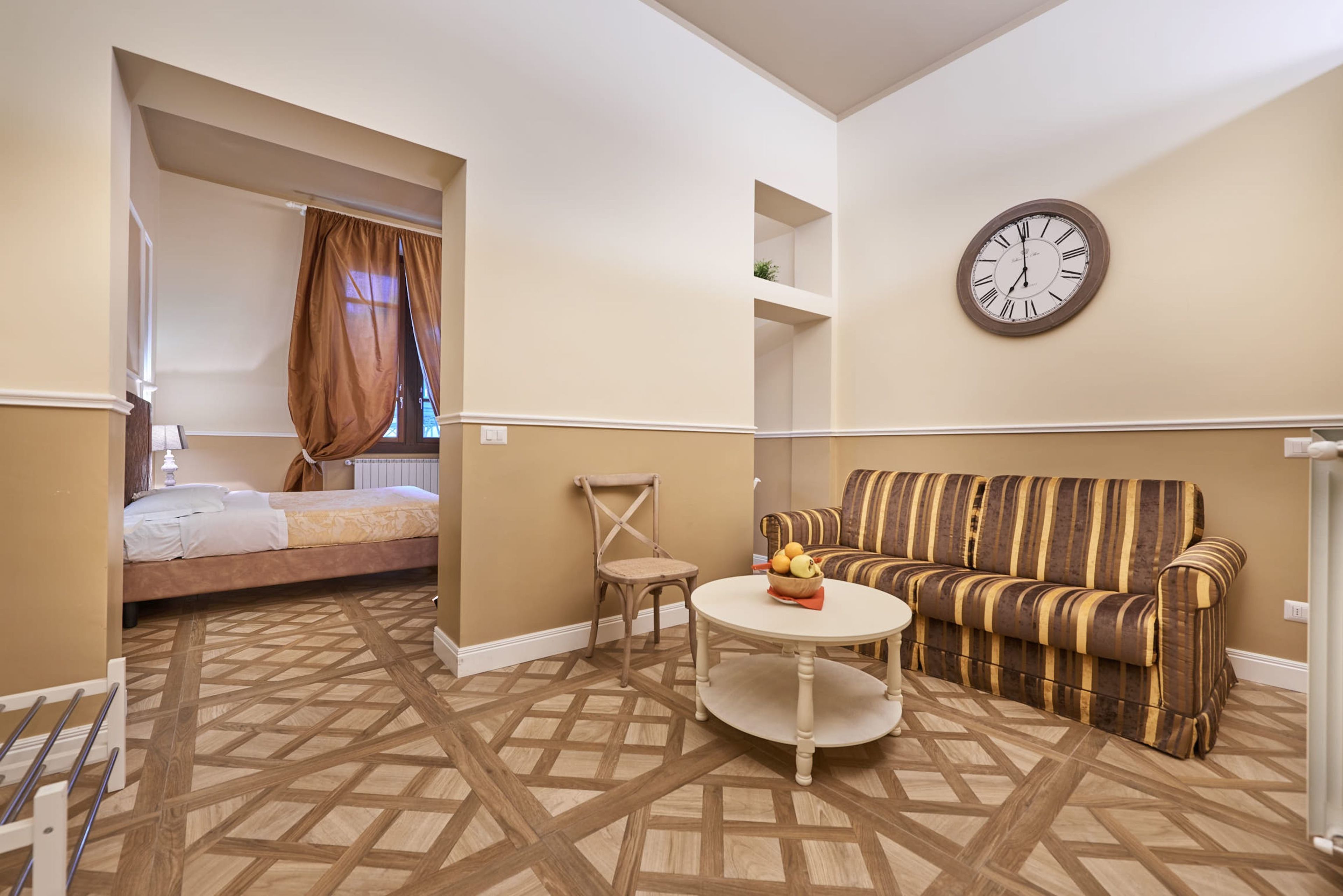 Una habitación de hotel con cama, sofá, una mesa redonda con fruta, un reloj en la pared y suelo estampado.