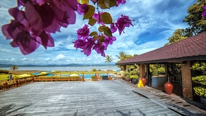 Terraza de madera con vista a un lago y montañas, zona de estar cubierta y plantas en macetas.