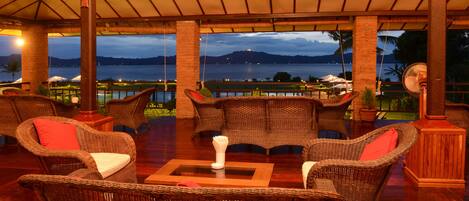 Un espacio lounge al aire libre con mobiliario de mimbre, una mesa central y vistas a un cuerpo de agua y montañas al fondo.