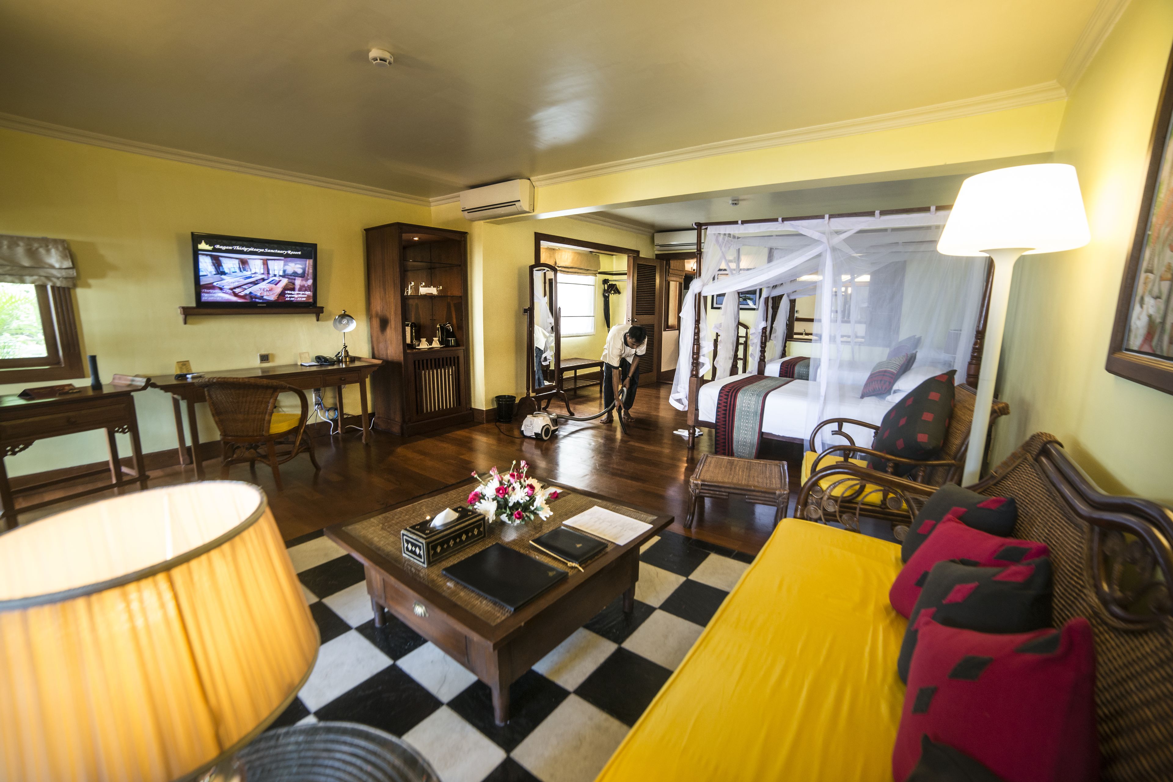 A hotel room with a checkered floor, a bed with a canopy, a television, a desk with a lamp, and a chair.