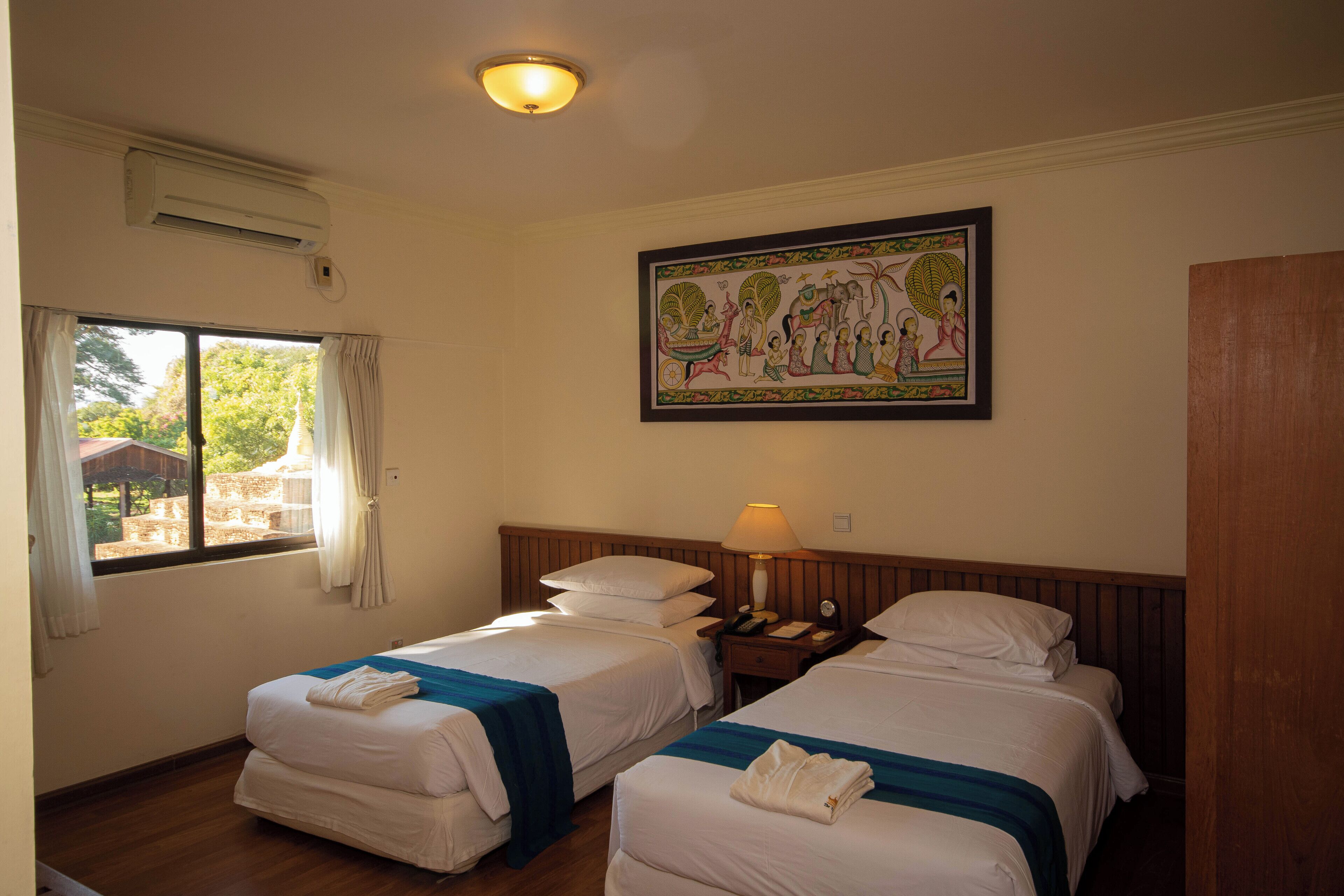 Habitación de hotel con dos camas, una ventana con cortinas, aire acondicionado de pared, mesita de noche con lámpara y un cuadro enmarcado sobre las camas.
