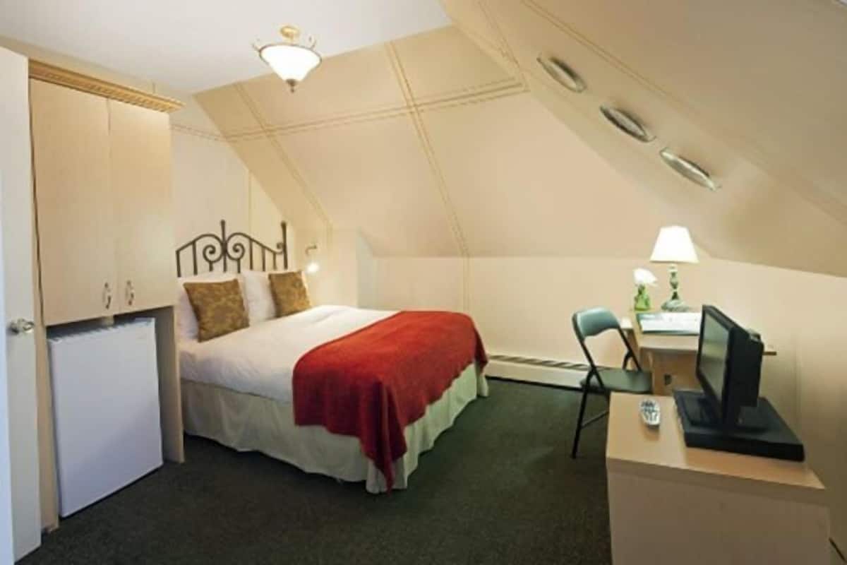 A small, single-room hotel room with a bed, desk, chair, and a small refrigerator.