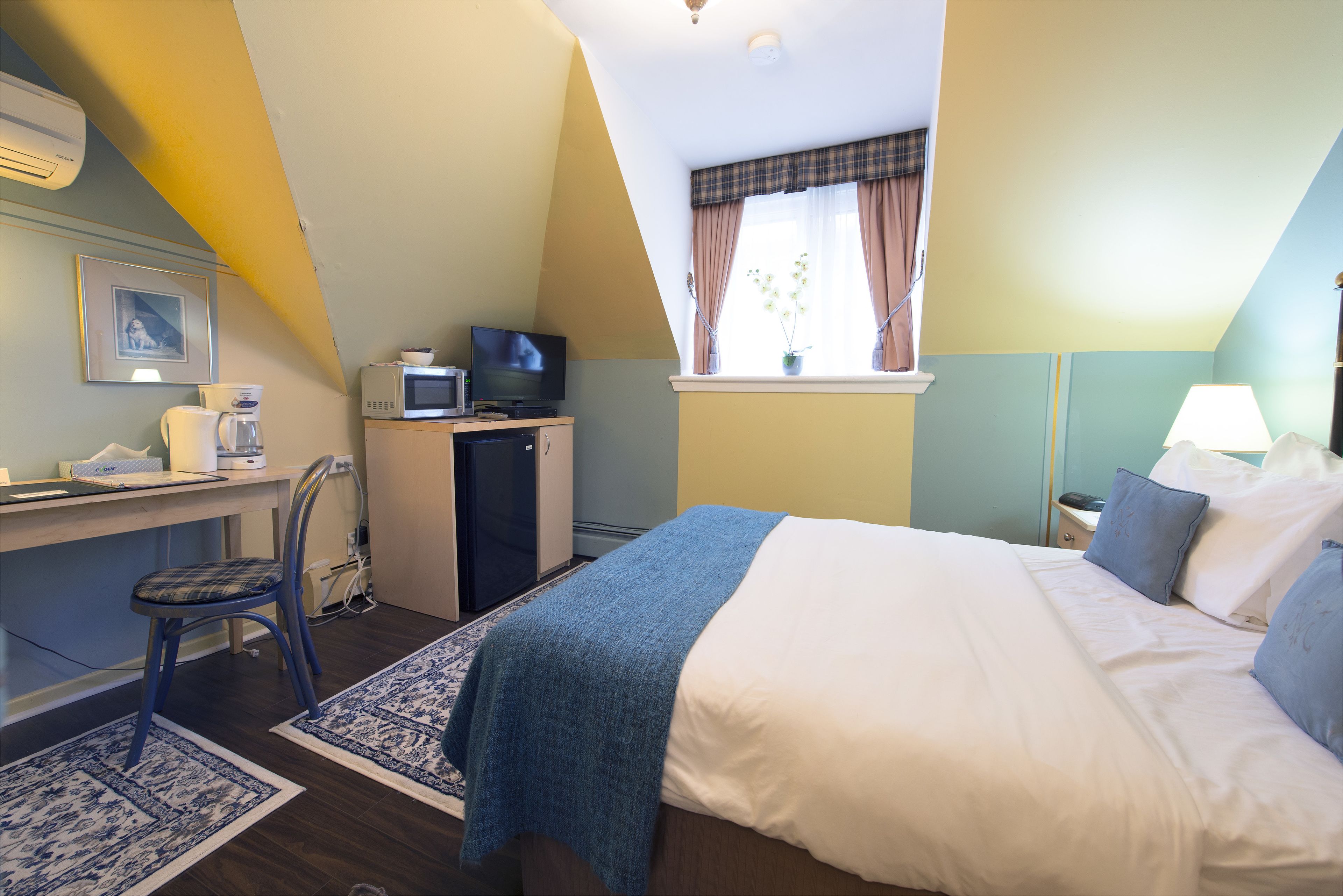 Quarto com cama, escrivaninha, cadeira, televisão e janela com cortinas.