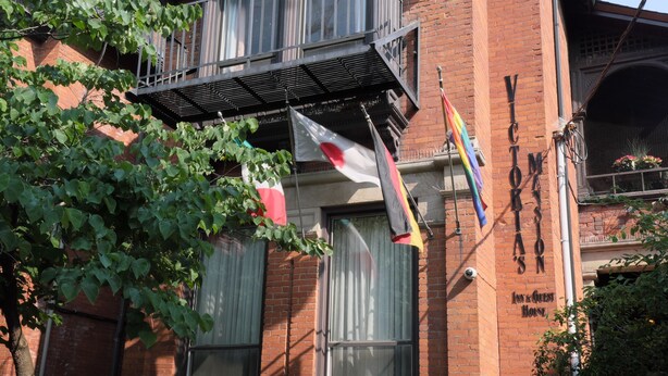 A two-story brick building with a balcony, a flag, and a sign that reads "VICTORIA'S". - Victoria's Mansion Guest House (Toronto)