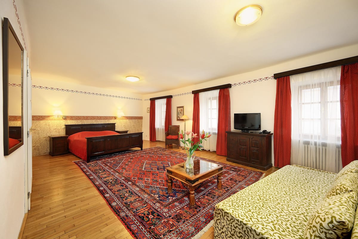 A hotel room with a bed, a sofa, a TV, a wooden coffee table, and a red patterned rug.