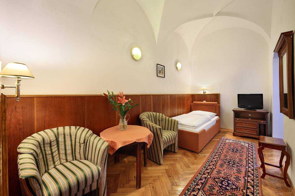 A room with a bed, two armchairs, a small table with a vase, a television, and a wooden cabinet.