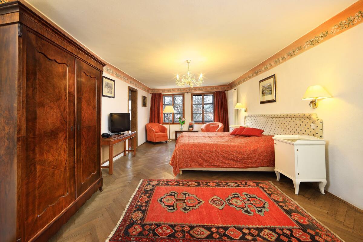 A bedroom with a wooden wardrobe, a television, a bed with red bedding, a nightstand, a lamp, and a window with curtains.