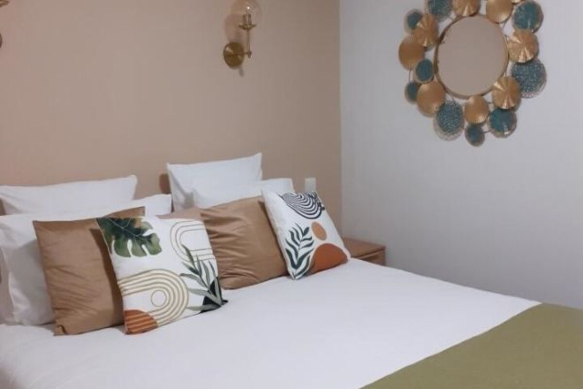 Una cama con varias almohadas, una lámpara fijada a la pared y un adorno colgante en la pared.