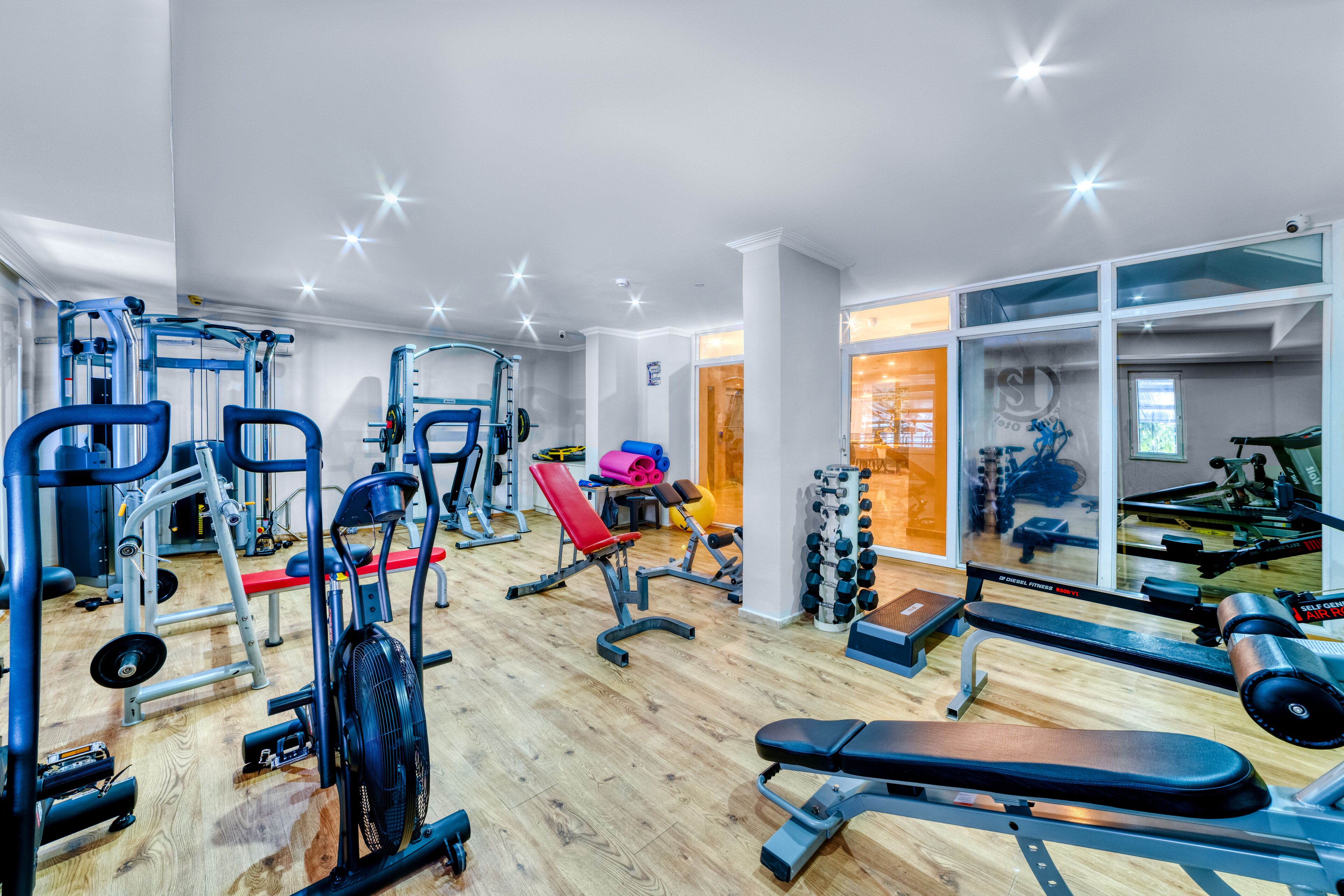 A gym with various exercise equipment, wooden flooring, and large windows.