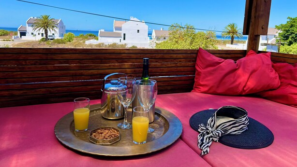 Une table avec un plateau de nourriture et de boissons, une bouteille et une paire de chaussures, le tout placé sur un balcon donnant sur une vue de la côte.
