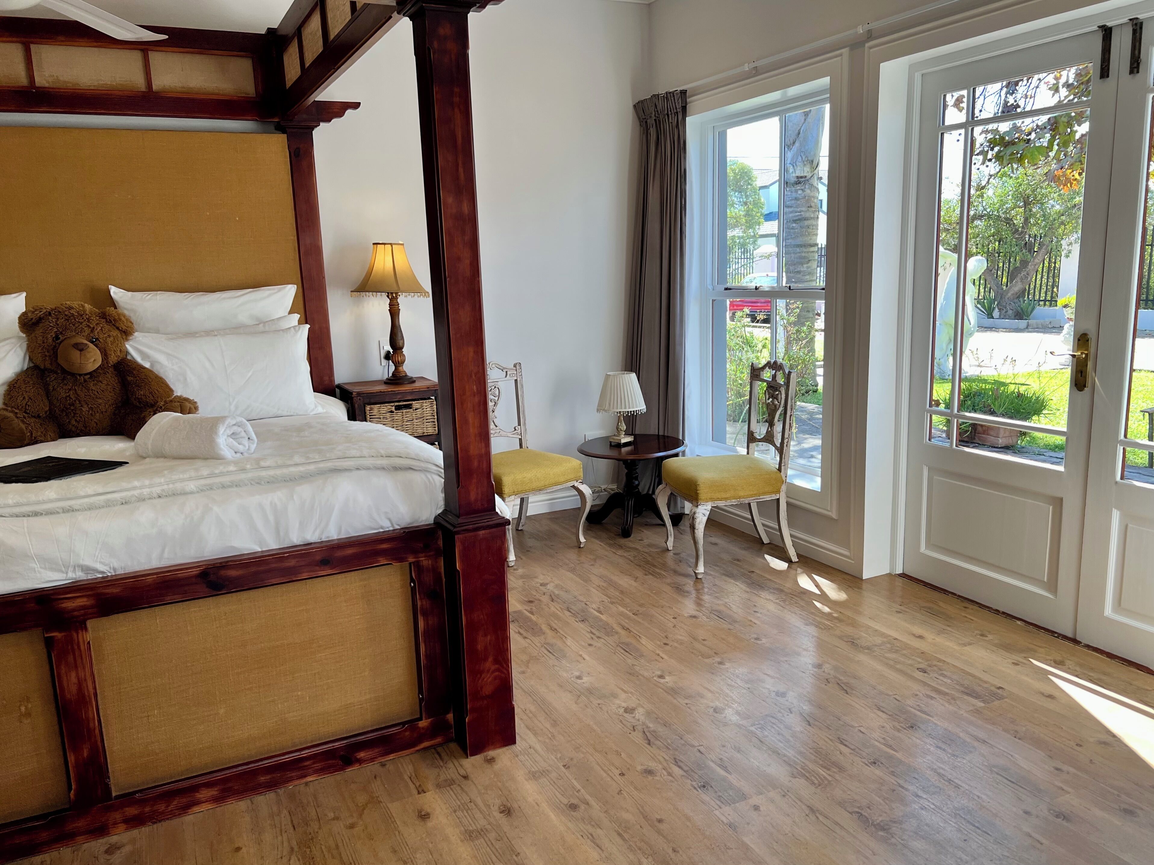 A four-poster bed with a teddy bear, a small table with a lamp, a chair, and a view through a glass door.