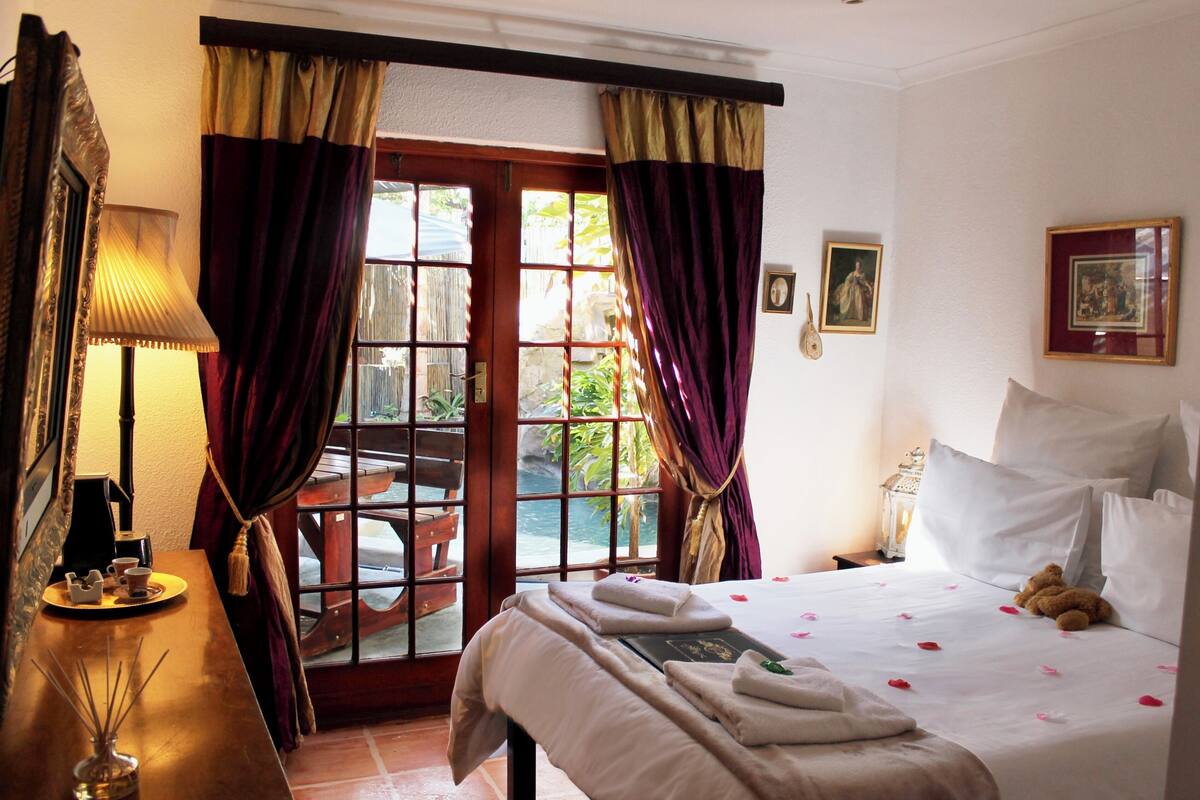 A hotel room with a bed, a bedside table, a lamp, a window with curtains, and a view of the outside.