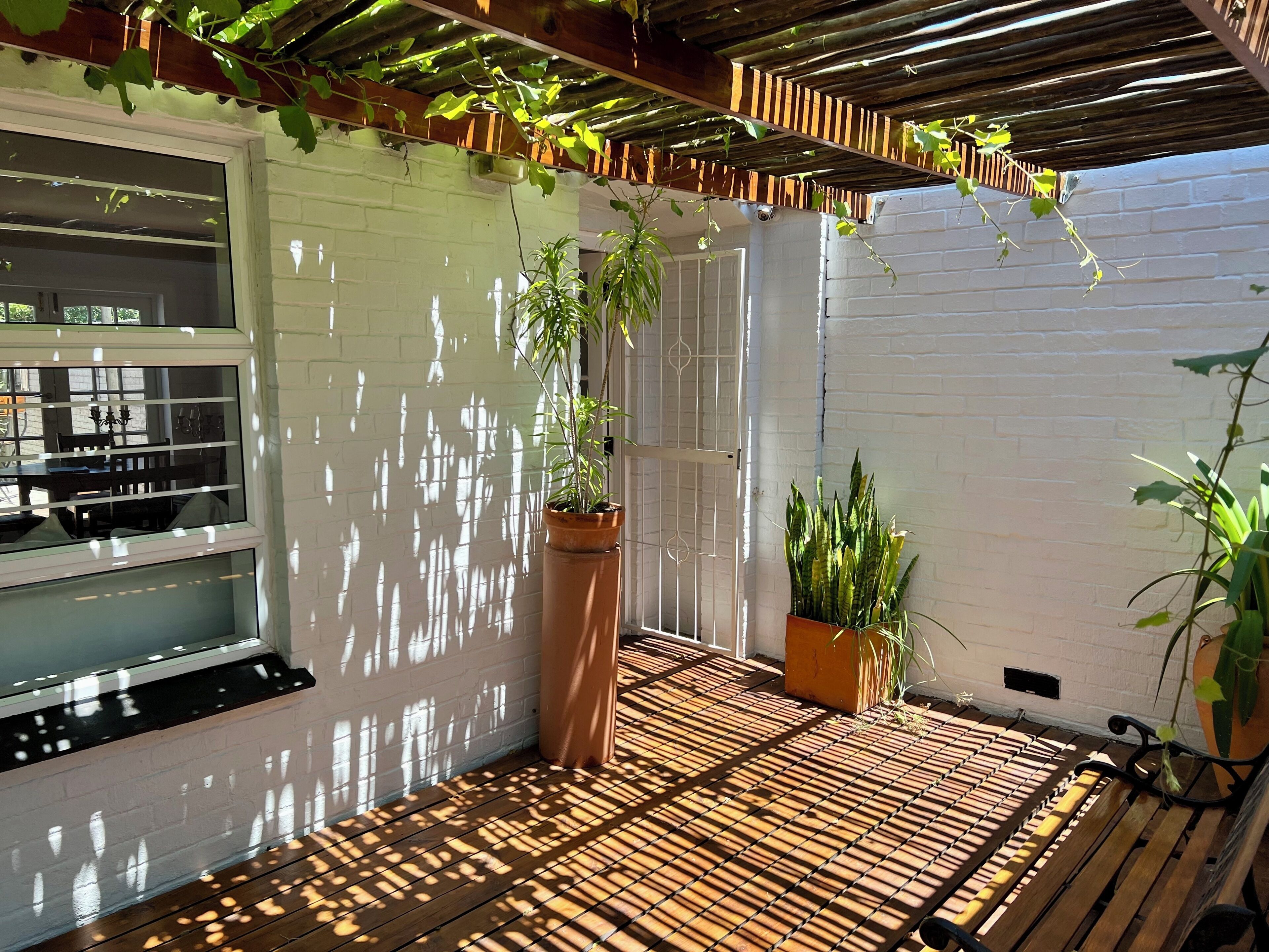 A covered patio with a wooden floor, potted plants, and a white brick wall.