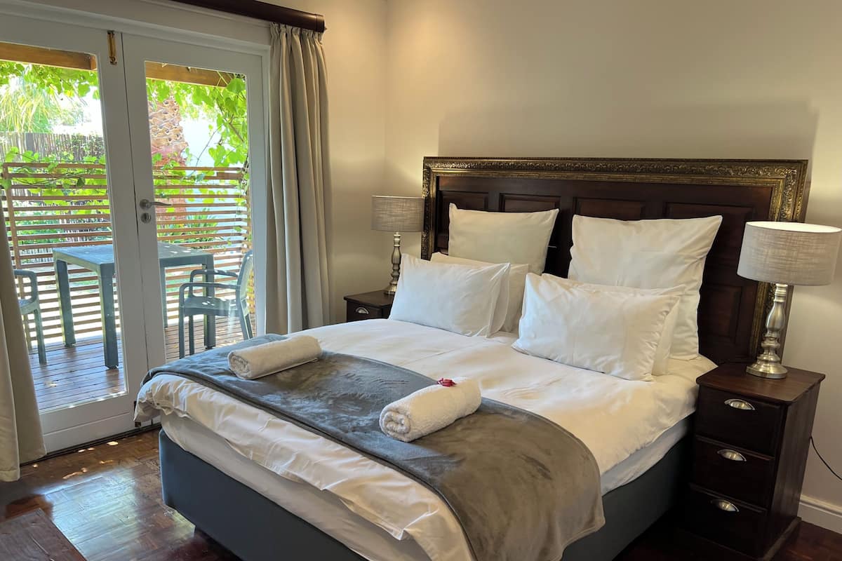 A neatly made bed with white linens, a grey throw blanket, and two towels. There is a wooden headboard, two bedside lamps, and a small wooden nightstand with drawers.