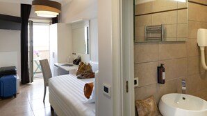 Una habitación de hotel con cama, escritorio, silla, maleta y baño con lavabo y espejo.