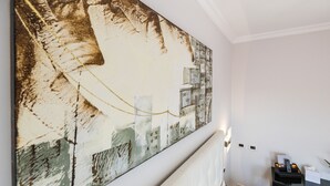 Habitación de hotel con un cuadro abstracto grande en la pared, una cama con ropa blanca, una mesita de noche con una lámpara y un pequeño escritorio con un teléfono y una caja de pañuelos.