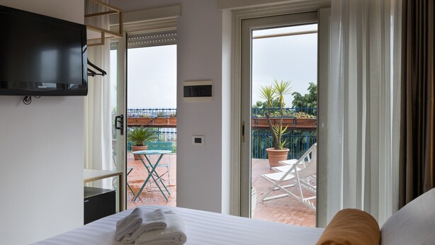 Habitación de hotel con cama, televisor montado en la pared y puerta corrediza de vidrio que da a un espacio exterior con mesa y sillas.