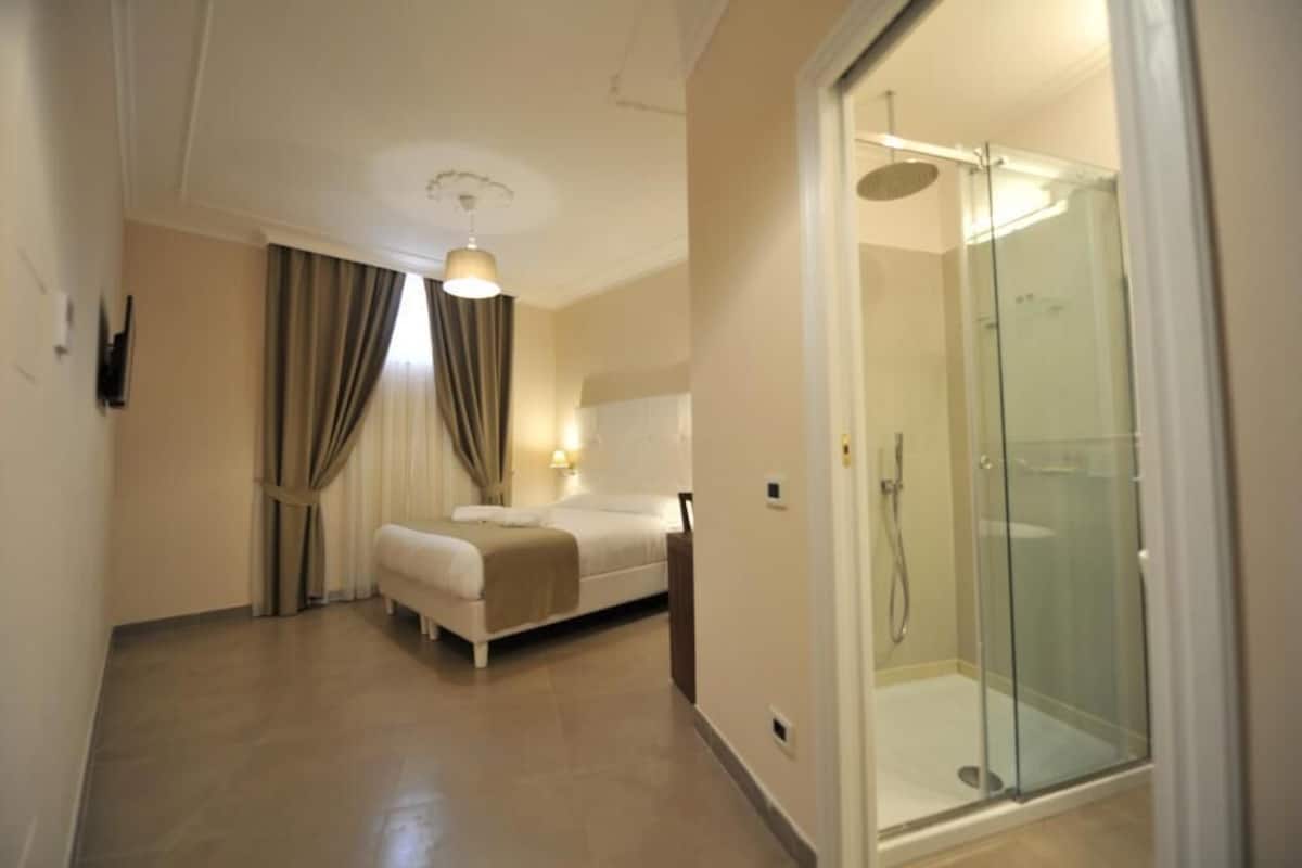 A hotel room with a bed, a glass-enclosed shower, and a ceiling light.