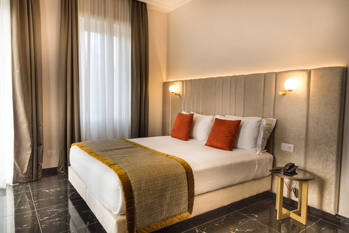 A hotel room with a large bed, a bedside table with a lamp, and a window with curtains.