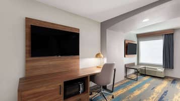 A hotel room with a wooden desk, a flat-screen TV, a lamp, a chair, a heater, and a large window with curtains.