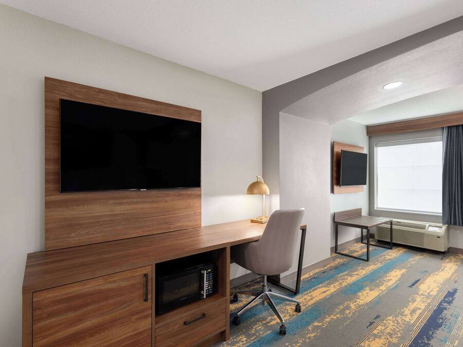 A hotel room with a wooden desk, a flat-screen TV, a lamp, a chair, a heater, and a large window with curtains.