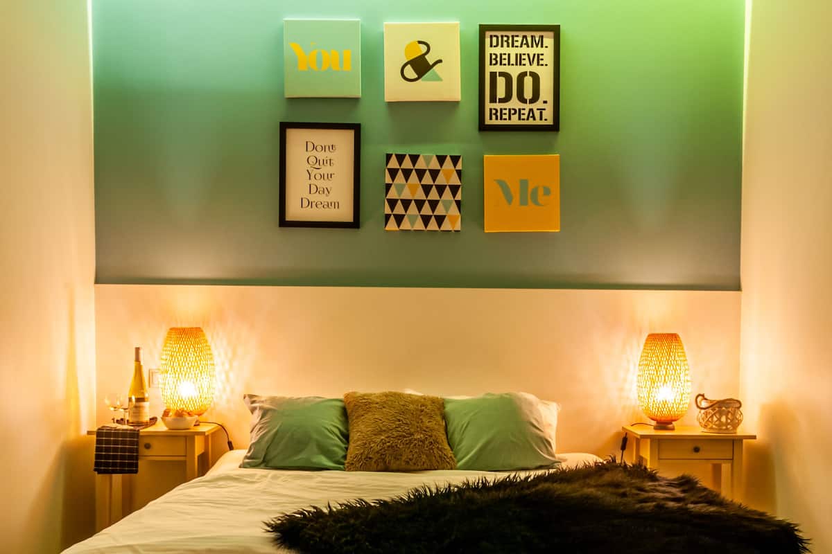 A bedroom with a bed, two bedside tables with lamps, a wall with motivational posters, and a wooden floor.