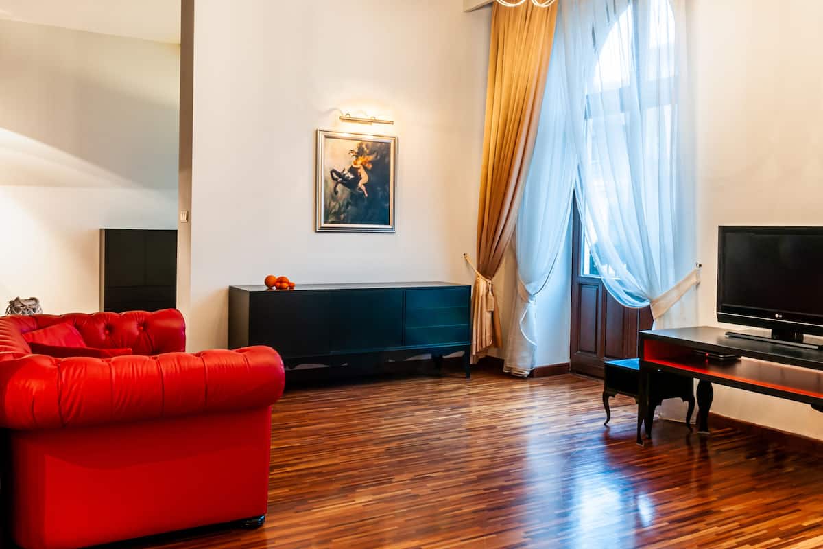 A living room with a red sofa, a TV on a stand, a painting on the wall, and a window with curtains.
