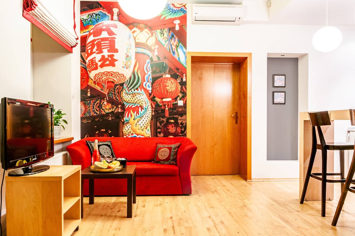 A living room with a red sofa, a TV on a wooden stand, a table with fruit, and a colorful mural on the wall.