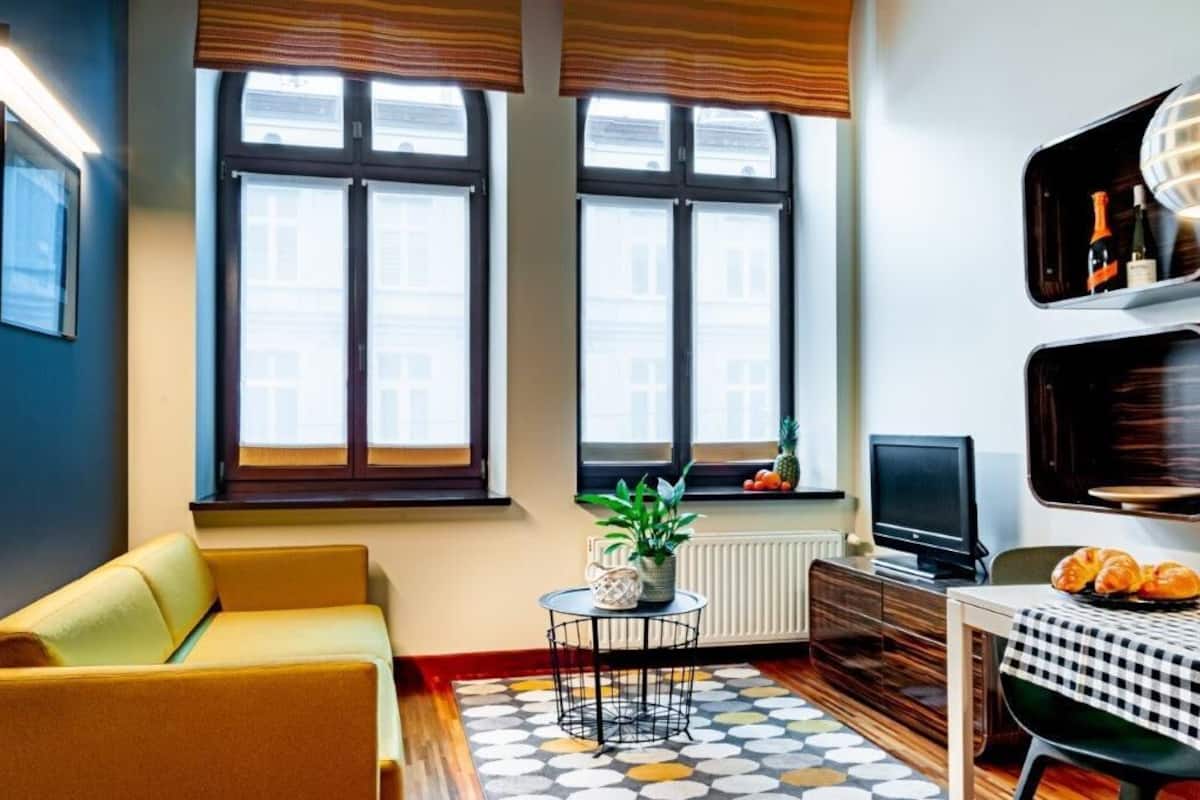 A cozy living room with a sofa, a small round table with a plant, a television, and a dining area with a checkered tablecloth.