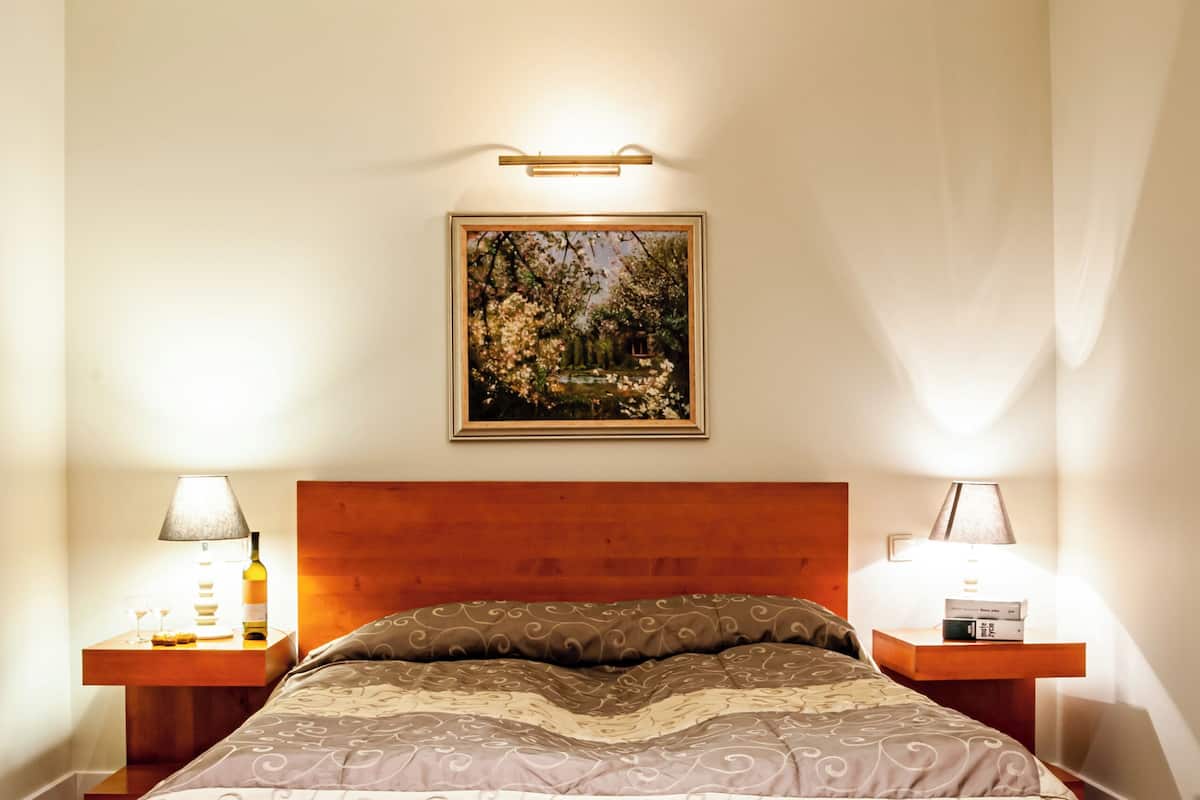 A bedroom with a wooden headboard, a bed with patterned bedding, two bedside tables with lamps, and a framed picture on the wall.