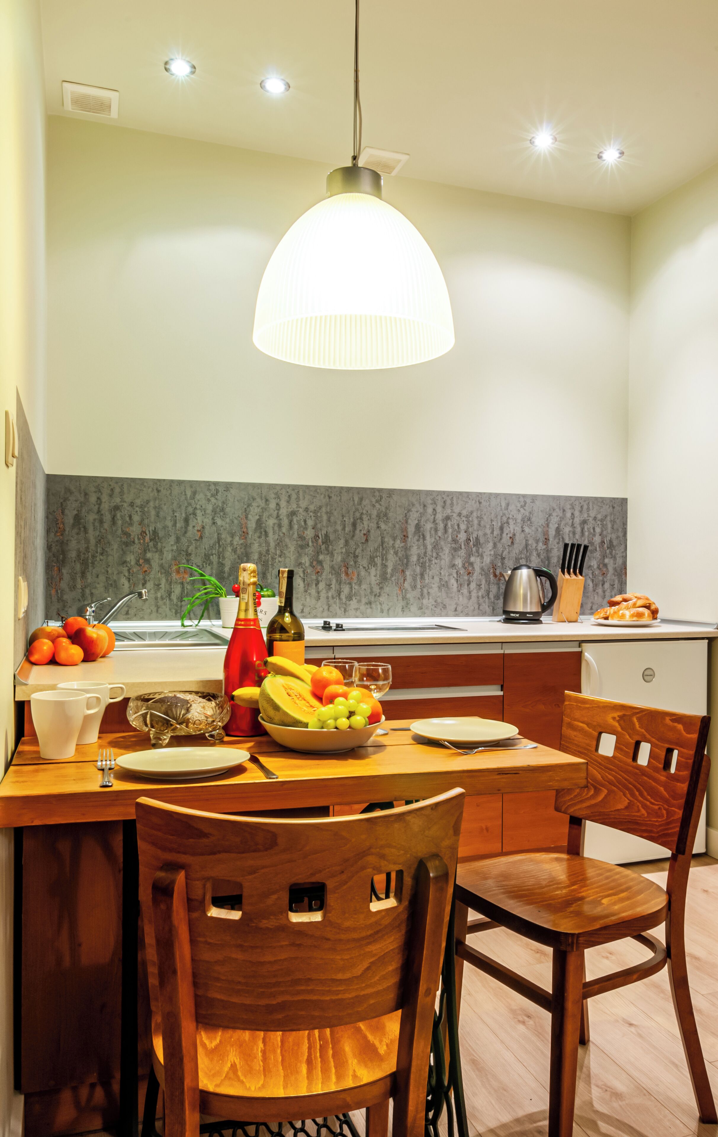 Une cuisine moderne avec une table à manger préparée pour deux personnes, agrémentée d’un panier de fruits, de bouteilles de vin et d’une machine à café.