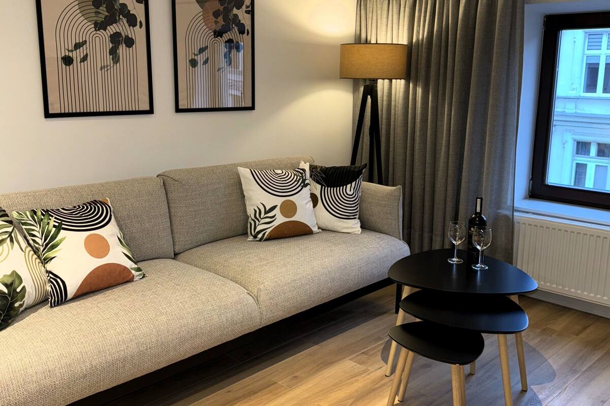 A modern living room with a sofa, two framed artworks, a floor lamp, a table with wine glasses, and a view of a building through a window with curtains.