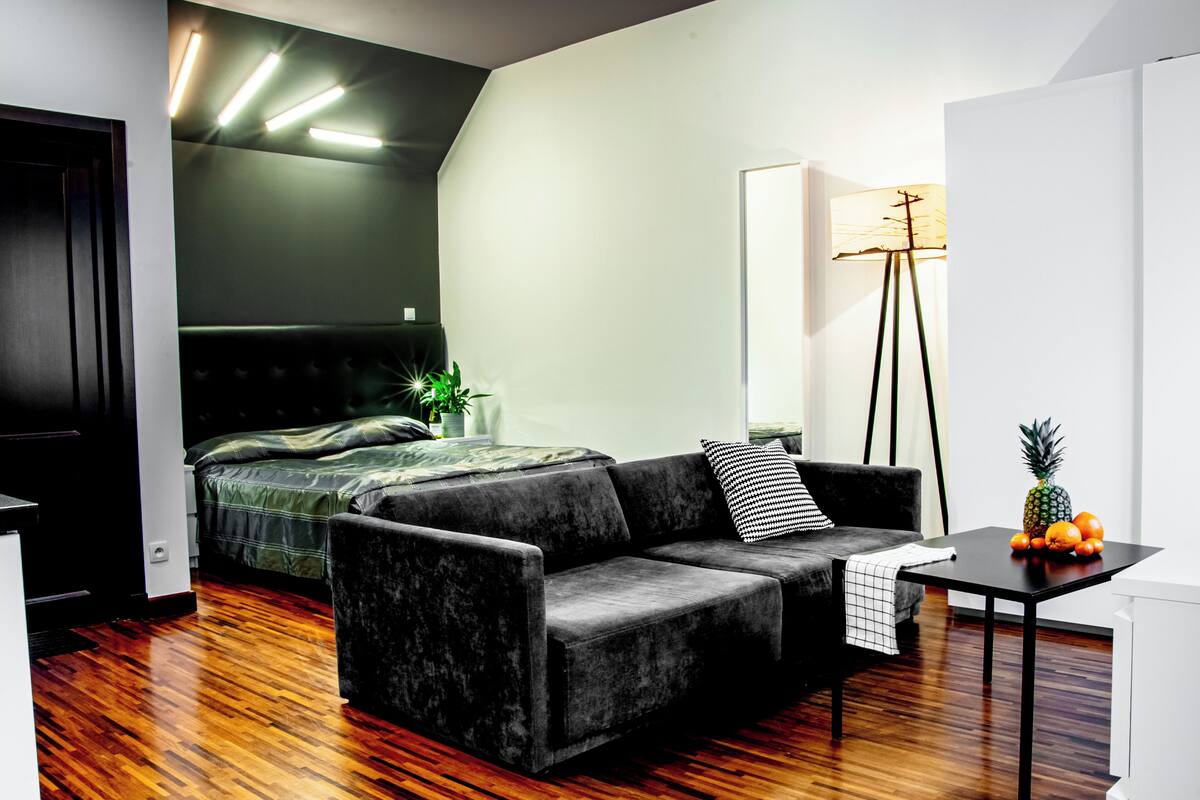 A modern hotel room with a dark green sofa, a small table with fruit, a bed with a green blanket, and a floor lamp.