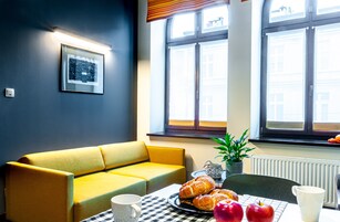 A cozy room with a yellow sofa, a table with pastries and apples, a potted plant, and a window with a view of buildings.
