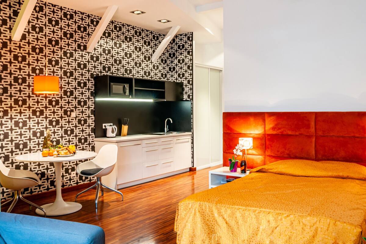 A modern hotel room with a kitchen area, a dining table, and a bed with a red headboard.