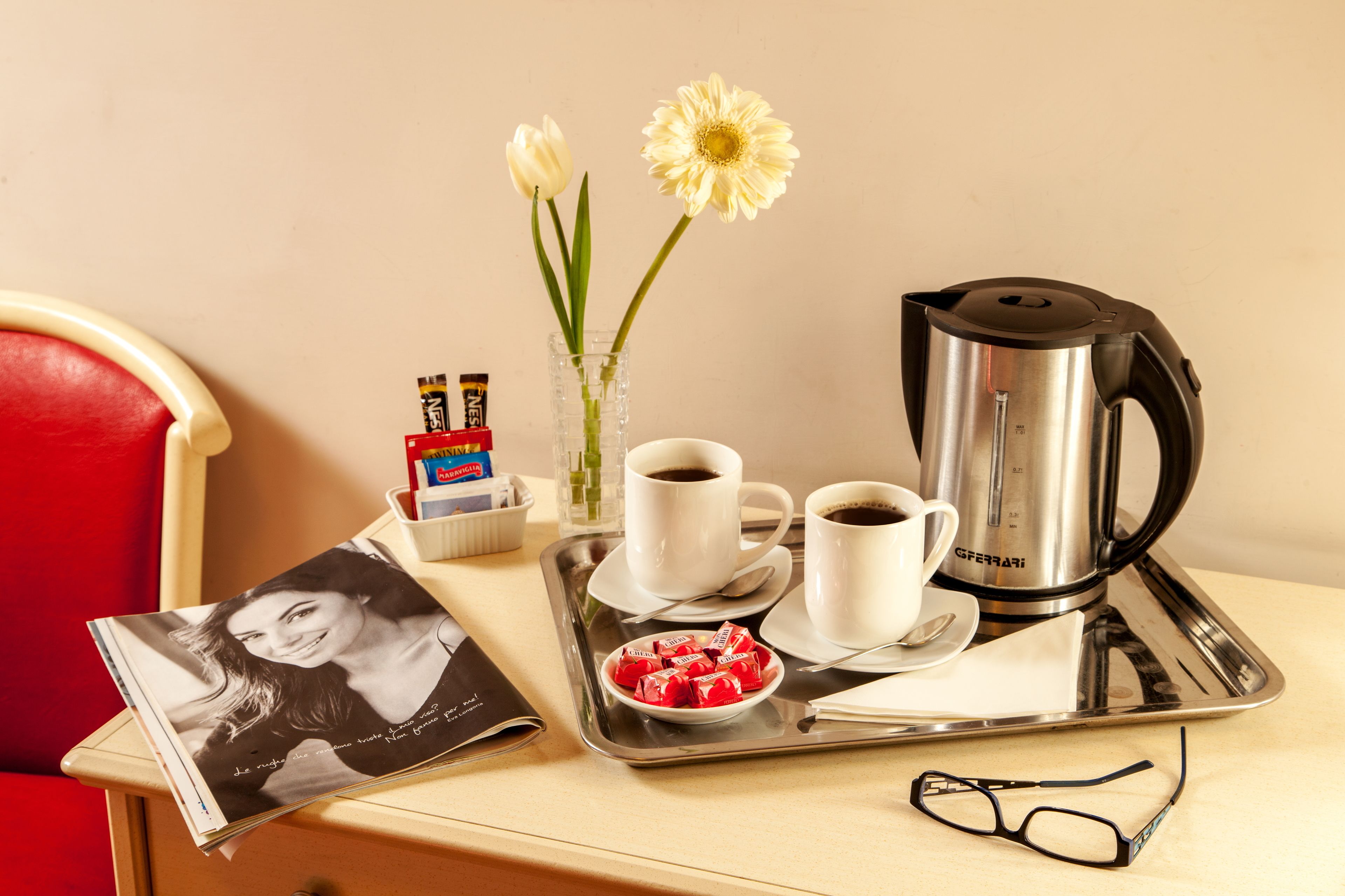 A tray with two cups of coffee, a coffee pot, a bowl of snacks, a magazine, a pair of glasses, and a vase with flowers.