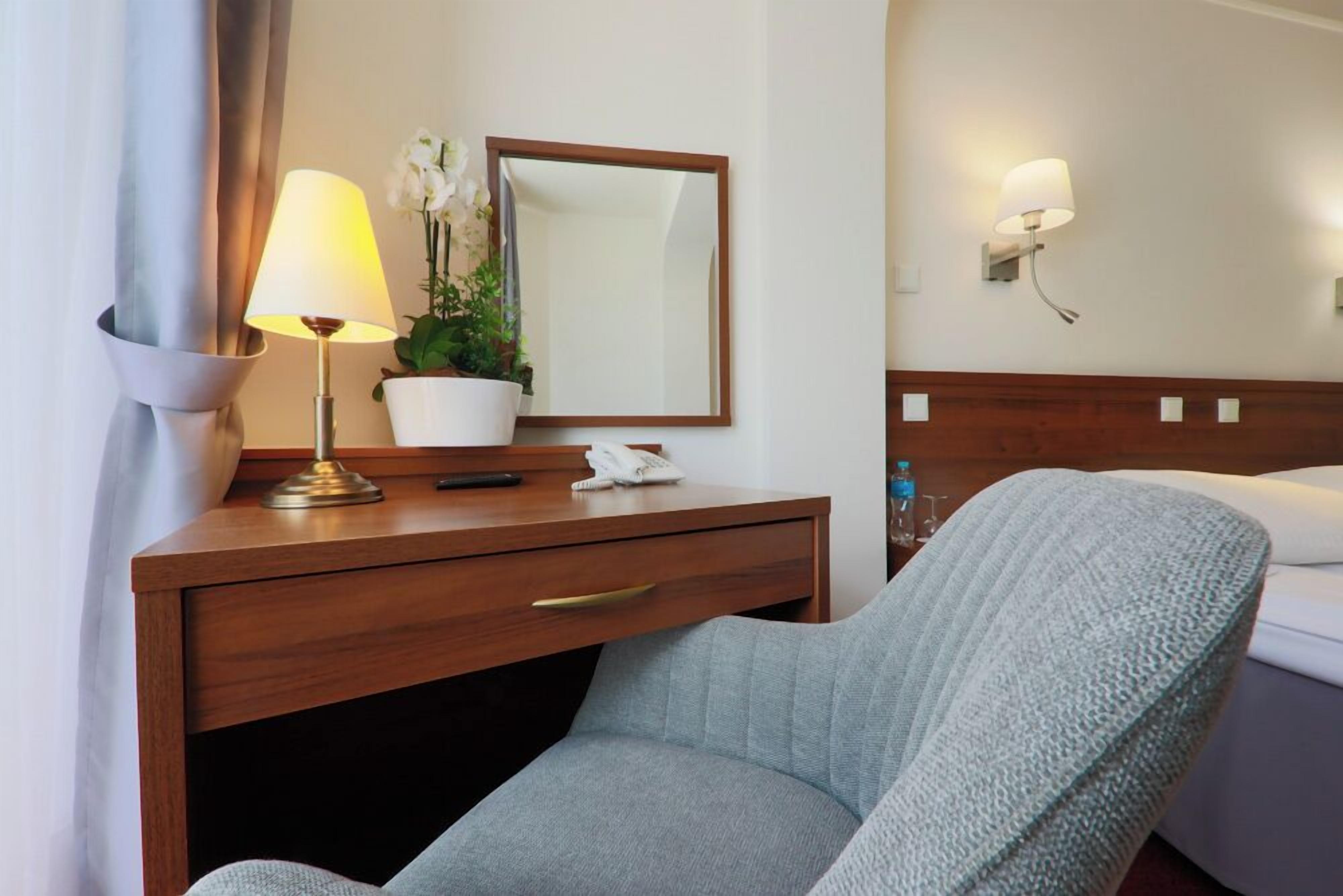Habitación de hotel con un escritorio de madera, una lámpara, un espejo, una planta en maceta, un teléfono y una cama con una colcha blanca.