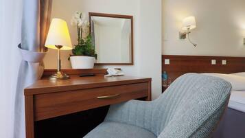 Habitación de hotel con un escritorio de madera, una lámpara, un espejo, una planta en maceta, un teléfono y una cama con una colcha blanca.