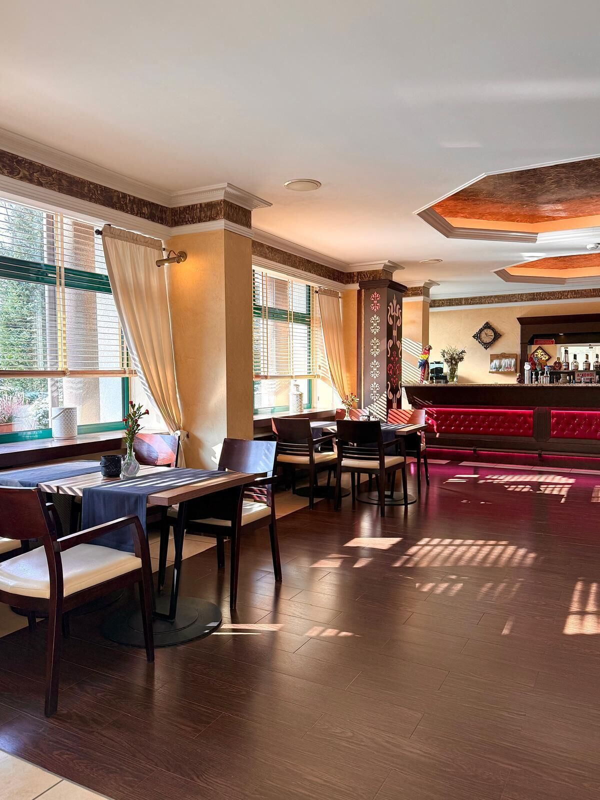 Un restaurant doté d’un plancher en bois, de tables avec des chaises, d’un bar et de grandes fenêtres ornées de rideaux.