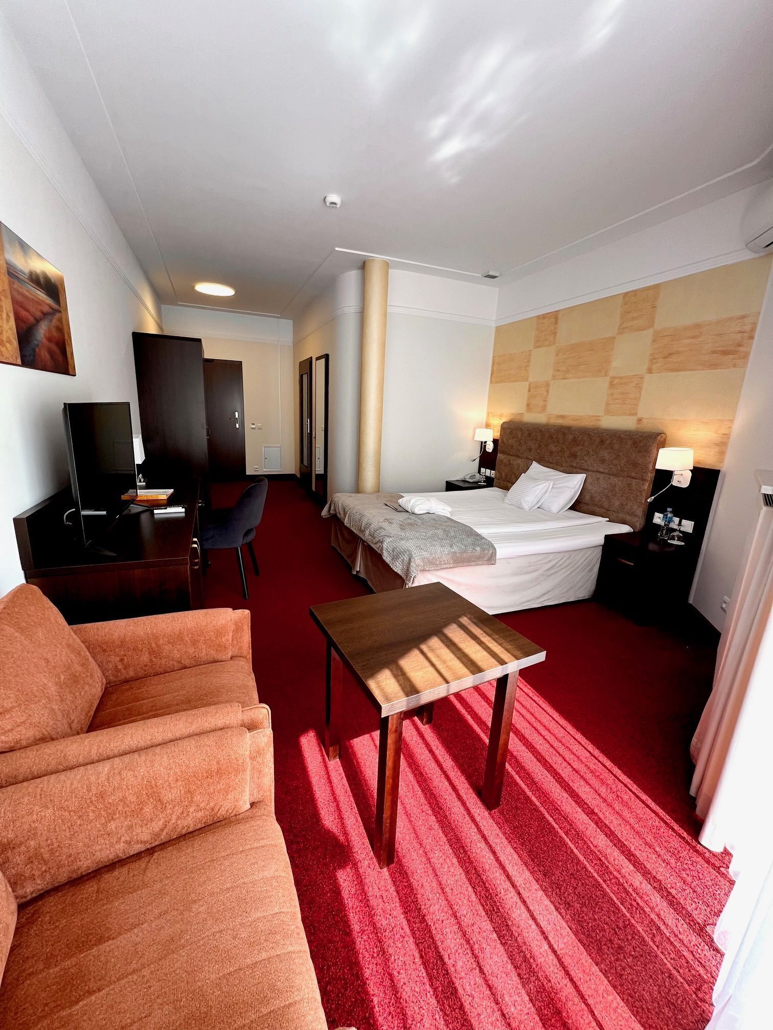 Quarto de hotel com cama, escrivaninha, cadeira, televisão e tapete vermelho.