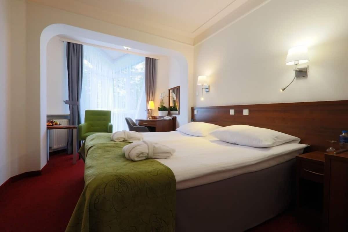 A hotel room with a large bed, a desk with a chair, a lamp, and a view through a window.