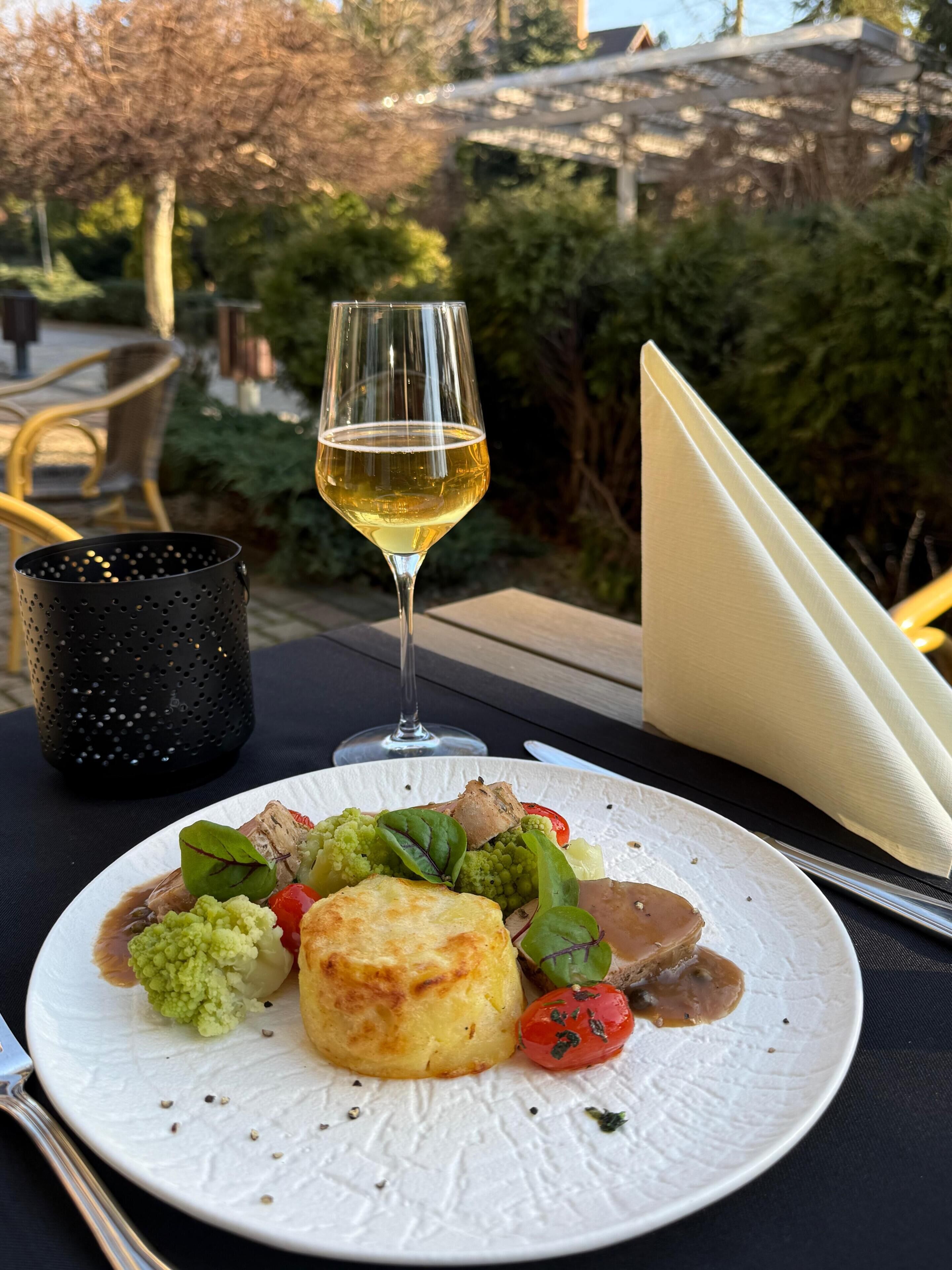 Une table à manger avec une assiette de nourriture, un verre de vin blanc et un récipient noir perforé.