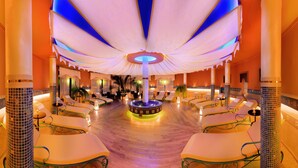 Un área de spa lujosa con múltiples tumbonas dispuestas alrededor de una piscina climatizada central, rodeada de columnas ornamentales e iluminación cálida.