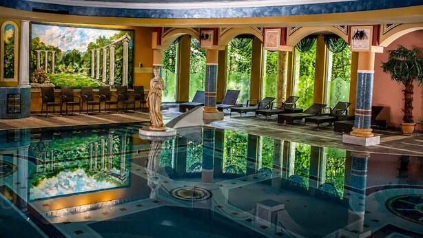 Un área de piscina con una estatua, sillas de descanso y un mural de columnas antiguas.