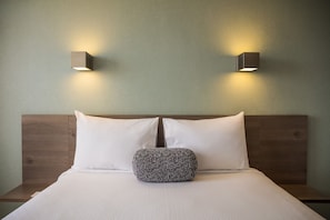 Un lit bien fait, avec du linge de lit blanc, un coussin à motifs et une tête de lit en bois. Deux lampes murales sont fixées de chaque côté du lit.