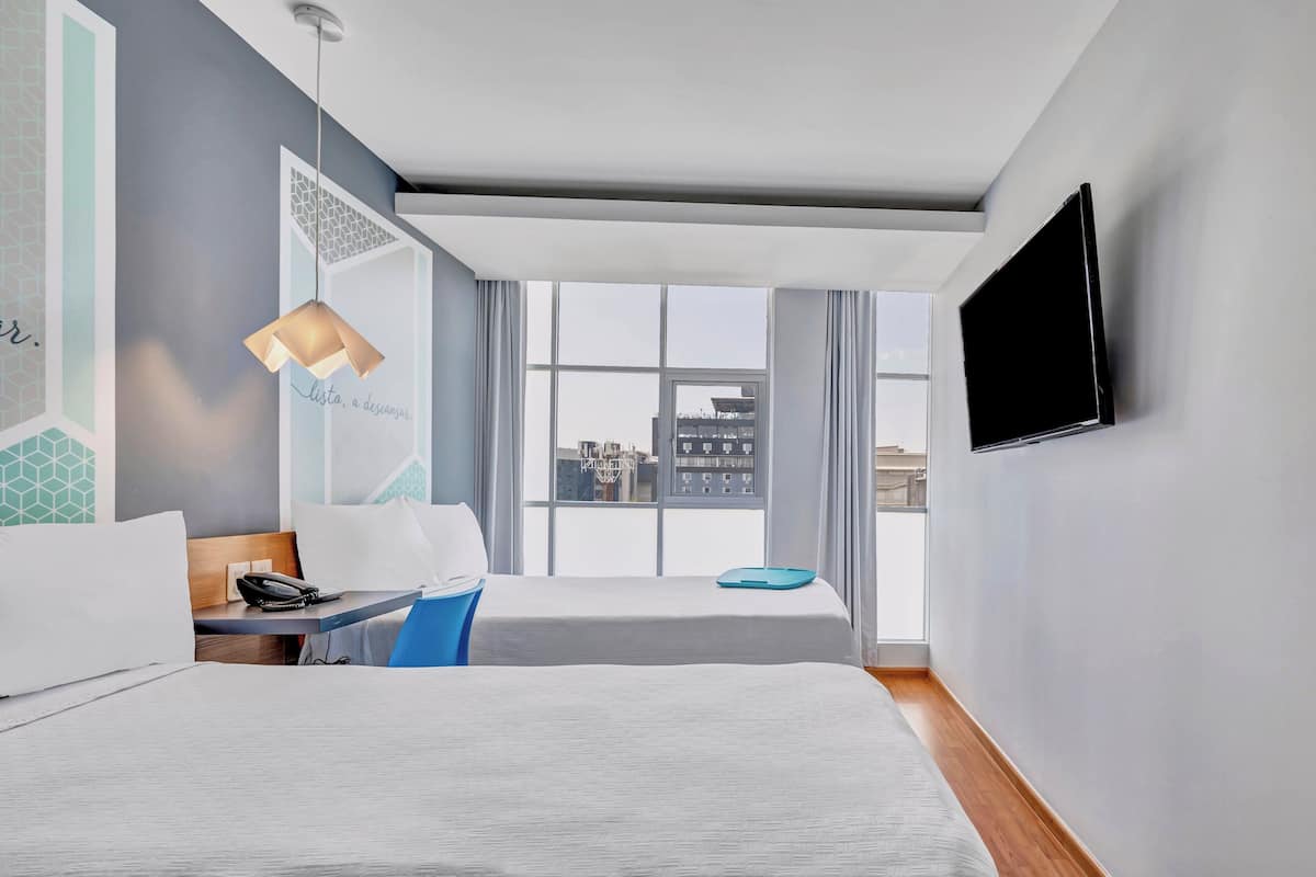 Ein modernes Hotelzimmer mit zwei Betten, einem Schreibtisch mit Telefon, einem blauen Sessel, einem an der Wand montierten Fernseher und großen Fenstern mit Blick auf die Stadt.
