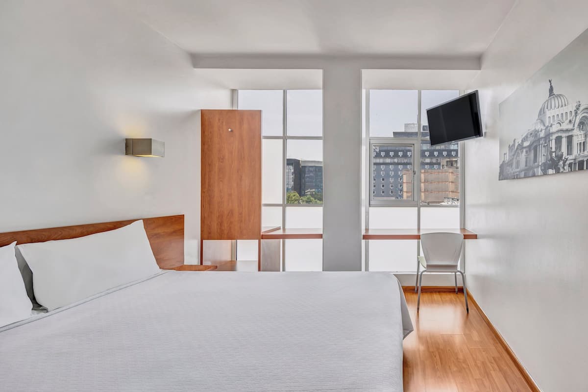 Ein modernes Hotelzimmer mit einem großen Bett, einem Holzschrank, einem Schreibtisch und einem Sessel. Ein Flachbildfernseher ist an der Wand befestigt, und durch die Fenster ist eine Stadtansicht sichtbar.