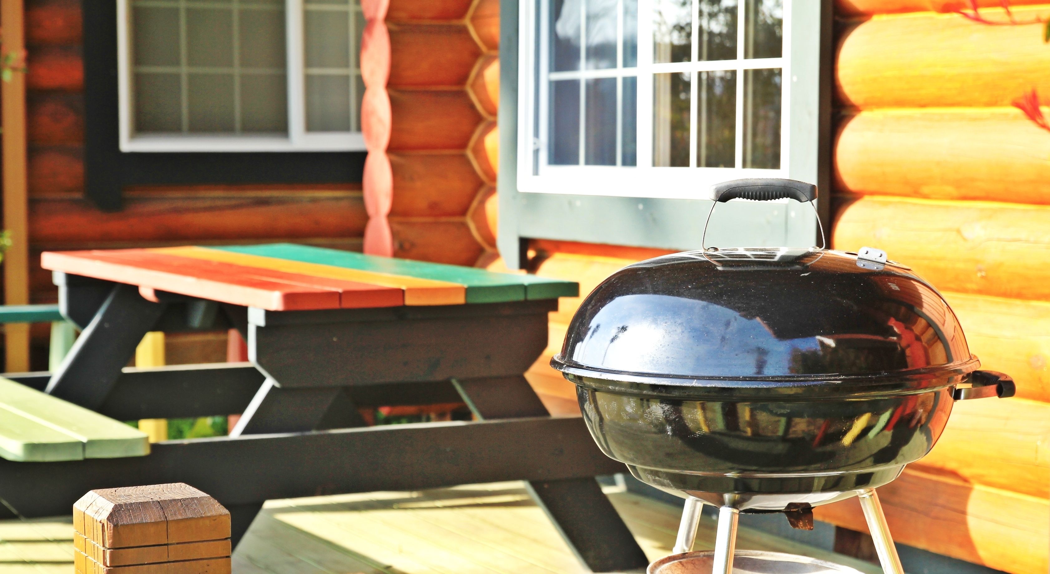 A barbecue grill in front of a wooden cabin with a window and a picnic table.